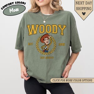 Puede incluir: Camiseta verde musgo con la palabra "WOODY" en letras amarillas en bloque sobre un gr&aacute;fico circular de un vaquero de dibujos animados. El gr&aacute;fico incluye el texto "EST 1995" y "TOY STOTY". La camiseta tiene mangas cortas y cuello redondo.