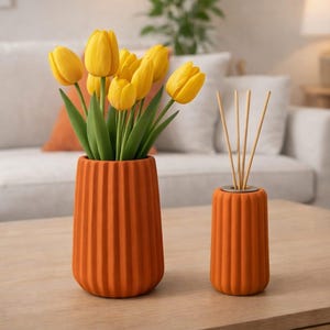 May include: An orange ceramic vase with vertical ridges holds yellow tulips. A smaller matching vase with reed diffuser sticks sits beside it. The vases are on a wooden table, with a sofa in the background.