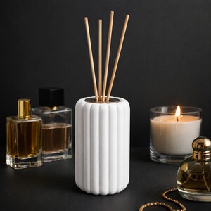May include: A white cylindrical reed diffuser with vertical fluted ridges and four light brown wooden reeds inserted into the top. The diffuser is surrounded by two glass perfume bottles, a lit white candle in a glass jar, and a gold chain.