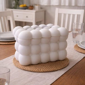 May include: A white, rectangular decorative box with a bubble-like design, resting on a woven placemat. The box is on a wooden table with a neutral-colored runner. A glass of water and a plate are also visible.