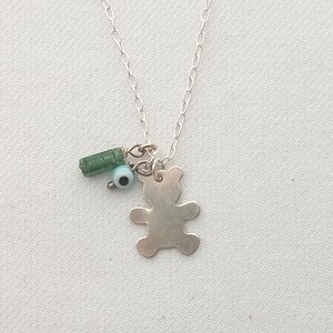 Puede incluir: Collar de plata con un colgante de oso de peluche, una cuenta cilíndrica verde y una pequeña cuenta azul con un centro negro. El collar cuelga sobre una superficie blanca.