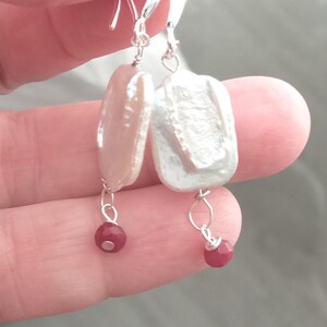 May include: A pair of elegant earrings featuring large, irregular-shaped, off-white pearls suspended from silver hooks. Each earring has a small, faceted, red gemstone dangling below the pearl, creating a striking contrast.
