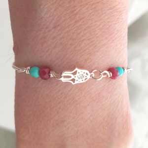 Op de afbeelding: Zilveren armband met een Hamsa-handbedel met een centrale edelsteen. De armband bevat turquoise en rode kralen. Het ontwerp heeft een fijne ketting, waardoor het een sieraad is.