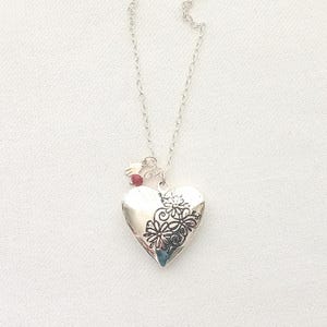 Puede incluir: Un colgante en forma de corazón de tono plateado suspendido de una delicada cadena de plata. El relicario presenta un diseño floral en relieve en el lado derecho y está adornado con una pequeña cuenta roja y un diminuto dije de tenedor.