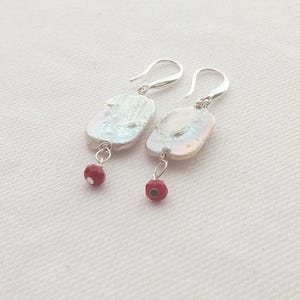 May include: A pair of silver earrings featuring large, irregular white pearls with a lustrous sheen. Each earring has a small, red bead dangling below the pearl. The earrings are displayed on a white surface.