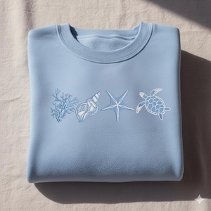 Pode incluir: Camisola azul clara com bordados com tema marinho. O design inclui um ramo de coral, uma concha, uma estrela do mar e uma tartaruga. A camisola está dobrada sobre uma superfície branca.