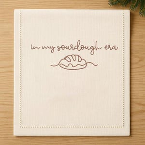 May include: Cream-colored linen kitchen towel with the phrase "in my sourdough era" and a bread loaf illustration in brown. The towel has a stitched border.