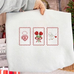 Op de afbeelding: Witte stof met drie rode borduurmotieven. De motieven omvatten een hartvormig medaillon, twee rozen met een strik en een duif met een hart. De motieven zijn omlijst met een rode geschulpte rand.