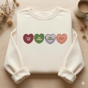Puede incluir: Sudadera color crema con cuatro caramelos bordados en forma de corazón. Cada caramelo tiene un color y mensaje diferente: rojo con "F*CK ICE", verde con "NO KINGS", morado con "PRO-CHOICE" y naranja con "FDT".