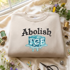 May include: A beige sweatshirt with the words "Abolish ICE" embroidered in black and a blue and white ice cube design. The sweatshirt is made of a soft material and is likely for casual wear.
