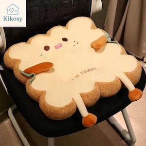 May include: A novelty cushion shaped like a slice of toast, with a smiling face and limbs. The cushion is tan with brown edges and the text "I'M TOAST". The arms and legs have orange details. It is placed on a black chair.