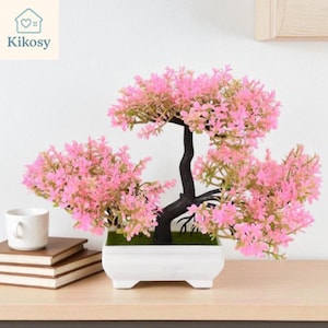 May include: A decorative bonsai tree with pink flowers and a dark brown trunk. The tree is in a white rectangular pot. The image also shows a white mug, a stack of books, and the text "Kikosy".