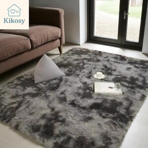 May include: A large, plush, gray area rug with a mottled pattern dominates the scene. A white triangular pillow rests on the rug near a brown sofa. A book and teacup sit on the rug, near a window.
