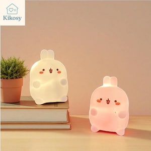 Pode incluir: Duas luzes noturnas em forma de coelho, uma branca e outra rosa, com traços fofos. As luzes estão brilhando, e a branca está em cima de uma pilha de livros. Uma pequena planta em vaso está à esquerda. A marca "Kikosy" está no canto superior esquerdo.