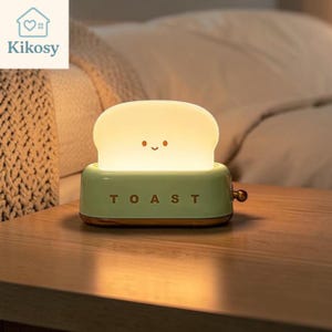 May include: A light green toaster-shaped night light with a glowing white bread slice insert featuring a simple smiling face. The word TOAST is printed in dark letters on the front, and a small round knob is located on the right side of the base.