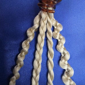 Op de afbeelding: Een haaraccessoire met een bruine schildpad-klauwclip die meerdere strengen gevlochten, lichtblond haar vasthoudt. De vlechten zijn strak geweven en hangen tegen een blauwe achtergrond.