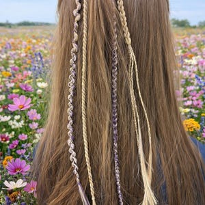 Op de afbeelding: Gevlochten haarverlengingen in tinten blond en paars zijn bevestigd aan lang, bruin haar. De haaraccessoires zijn gestyled met een natuurlijke, bohemien uitstraling. De achtergrond toont een levendig veld met wilde bloemen onder een blauwe lucht.