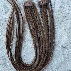 May include: A collection of brown hair extensions with various braid styles. The extensions include clip-in pieces and loose strands, showcasing different shades of brown. The hair is arranged on a white, textured surface.