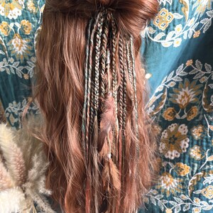 May include: Reddish-brown hair styled in a bun, adorned with multiple braided hair wraps. The wraps feature strands of green, brown, and cream-colored threads, accented with small feathers. The background is a teal fabric with a floral pattern.