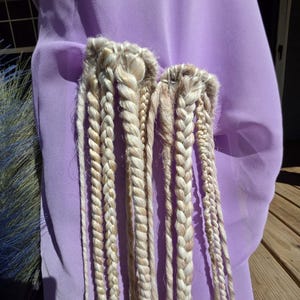 May include: Close-up of two sets of blonde braided hair extensions. The braids are thick and hang down, with a light brown hue. The background is a solid lavender fabric.