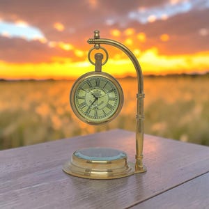May include: A brass pocket watch-style clock with a cream-colored face and Roman numerals. The clock is suspended from a curved stand and sits on a round base. The clock face reads "VICTORIA LONDON 1876".