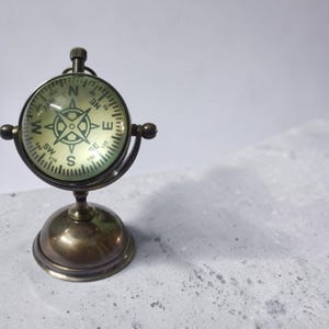 May include: A vintage-style brass compass with a glass face, mounted on a round base. The compass dial features directional markings and a central star design. The compass is approximately 4 inches tall.
