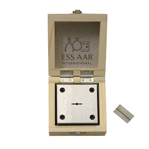 Op de afbeelding: Open houten kist met een metalen matrijs en een klein metalen stuk. De kist heeft de tekst "ESS AAR INTERNATIONAL" en een logo. De matrijs heeft een kleine, horizontale sleuf. De kist is open en toont de inhoud.