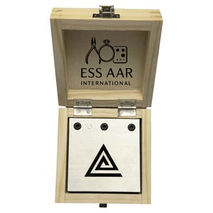 Pode incluir: Caixa de madeira aberta com tampa articulada. No interior, uma placa de metal apresenta um design geométrico preto. A caixa é marcada com o texto "ESS AAR INTERNATIONAL" e um logotipo de ferramentas de joalheria.