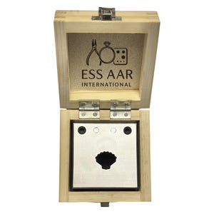 Pode incluir: Caixa de madeira aberta com tampa articulada, revelando uma placa de metal com um recorte em forma de concha. A caixa é marcada com o texto "ESS AAR INTERNATIONAL" e um logotipo de ferramentas de joalheria.