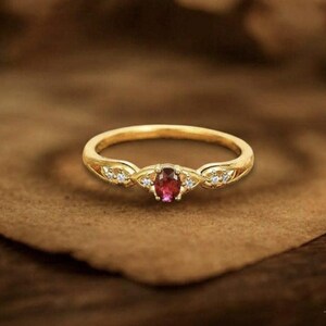 Könnte beinhalten: Ein goldener Ring mit einem ovalen, roten Edelstein in der Mitte. Der Ring hat kleine, klare Edelsteine auf beiden Seiten des zentralen Steins. Der Ring liegt auf einer braunen Oberfläche.