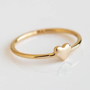 May include: A delicate gold ring with a small heart-shaped charm. The ring has a thin band and a polished finish, reflecting light. The heart charm is centered on the ring, creating a simple yet elegant design.