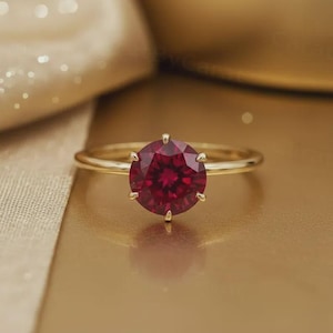 May include: A gold ring featuring a large, round, deep red gemstone. The gemstone is the focal point, set with six prongs. The ring band is a simple, polished gold. The background is a warm, golden hue.