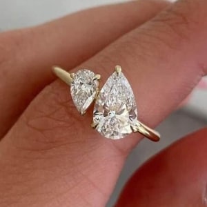 May include: A gold ring featuring two pear-shaped, clear gemstones. The larger gemstone is centered, while the smaller one is positioned to the side. The ring's band is a simple, polished gold. The gemstones sparkle, reflecting light.