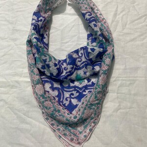 May include: A blue, white, and teal paisley patterned scarf. The scarf has a triangular shape and is made of a lightweight fabric. The edges are trimmed with a floral border in shades of pink and teal.