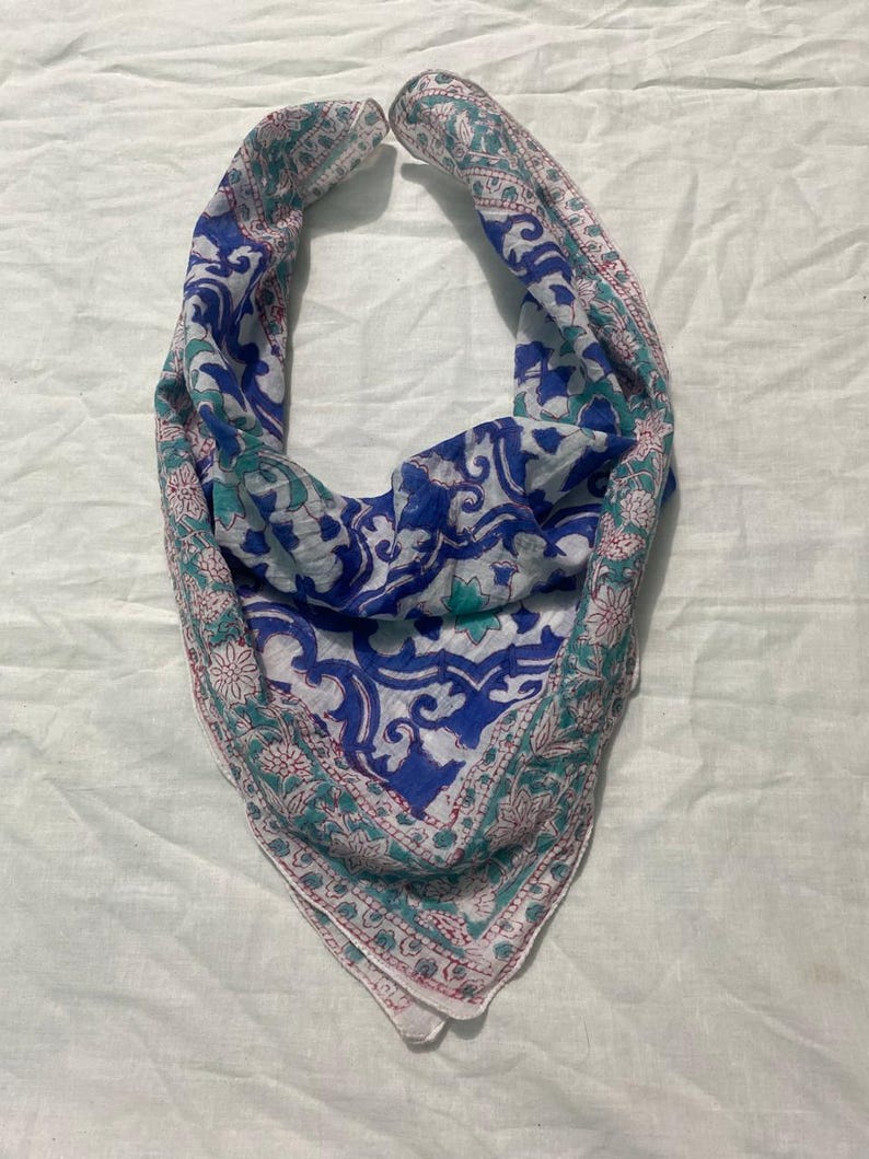 May include: A blue, white, and teal paisley patterned scarf. The scarf has a triangular shape and is made of a lightweight fabric. The edges are trimmed with a floral border in shades of pink and teal.