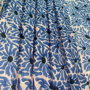 Könnte beinhalten: Ein Stoff mit einem sich wiederholenden Blumenmuster. Das Design zeigt große blaue Blumen mit schwarzen Zentren vor einem hellrosa Hintergrund. Der Stoff ist gefaltet, wodurch ein strukturierter Eindruck entsteht.