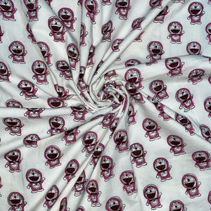 Puede incluir: Tela blanca con un patrón repetido de un personaje de dibujos animados en rosa y granate. El personaje tiene una cabeza grande, una campana y una bolsa. La tela es probablemente de algodón o similar.