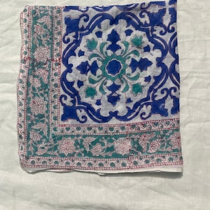May include: A folded square textile with a white background and a blue, teal, and pink floral pattern. The center features a large blue and teal floral design, while the borders have a repeating floral pattern in teal and pink.