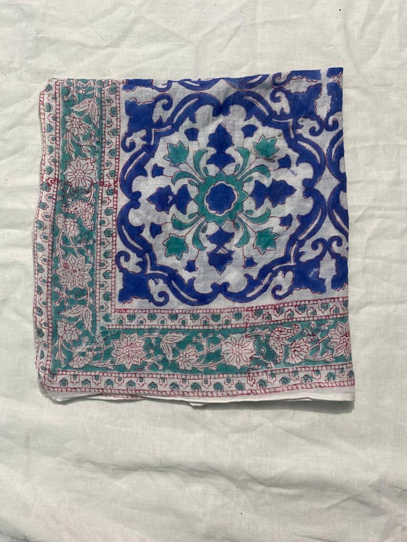 May include: A folded square textile with a white background and a blue, teal, and pink floral pattern. The center features a large blue and teal floral design, while the borders have a repeating floral pattern in teal and pink.