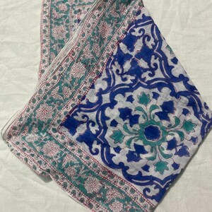 May include: A white scarf with a blue and teal floral pattern. The scarf has a border with a repeating floral design in teal and pink. The center of the scarf features a large blue floral design.