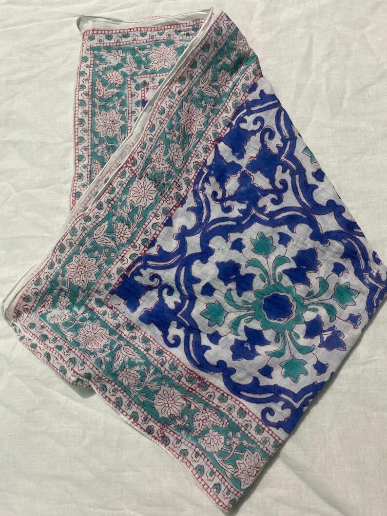 May include: A white scarf with a blue and teal floral pattern. The scarf has a border with a repeating floral design in teal and pink. The center of the scarf features a large blue floral design.