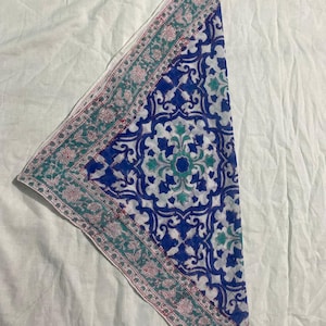 May include: A folded, patterned textile with a white background. The textile features a central design of blue and white floral patterns. A border of teal and pink floral designs frames the central pattern. The textile is likely a scarf or bandana.