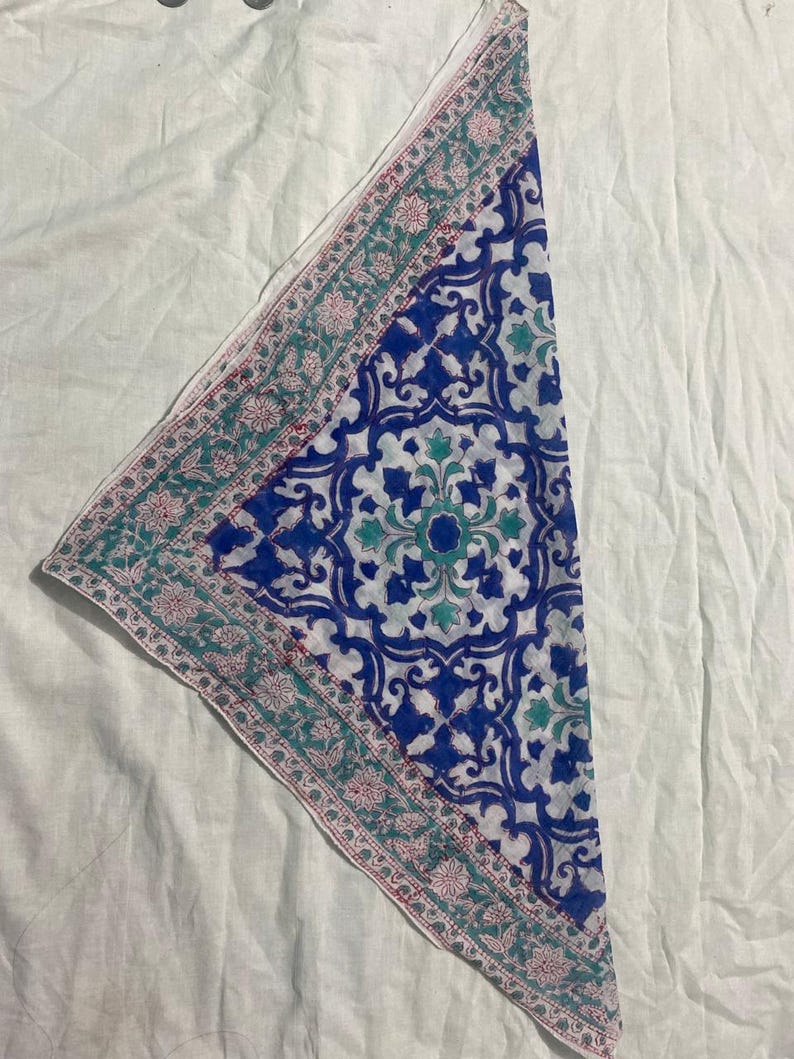 May include: A folded, patterned textile with a white background. The textile features a central design of blue and white floral patterns. A border of teal and pink floral designs frames the central pattern. The textile is likely a scarf or bandana.