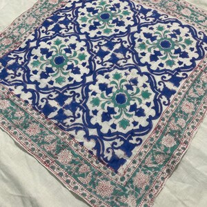 May include: A square, white fabric with a detailed, symmetrical pattern in blue, teal, and red. The central design features floral and geometric shapes, framed by a border of floral motifs. The fabric is likely a decorative textile, such as a scarf or a small tablecloth.