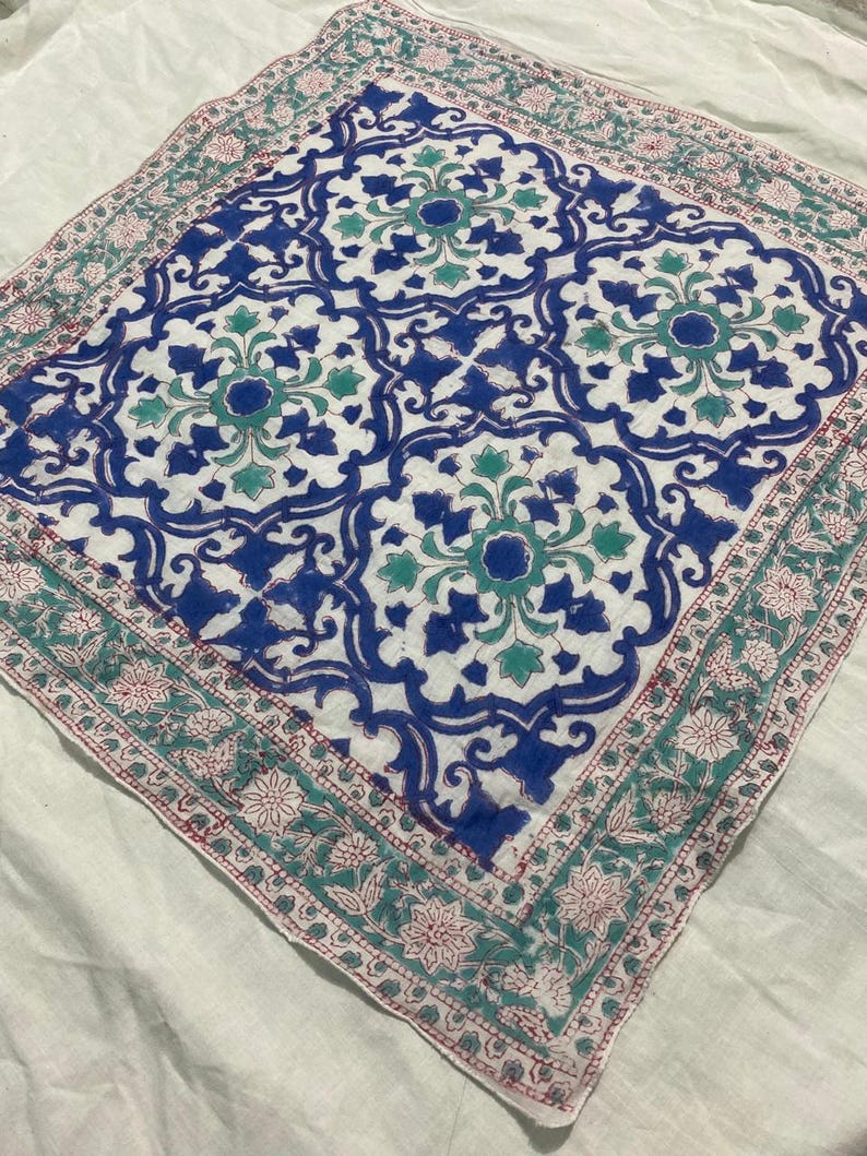 May include: A square, white fabric with a detailed, symmetrical pattern in blue, teal, and red. The central design features floral and geometric shapes, framed by a border of floral motifs. The fabric is likely a decorative textile, such as a scarf or a small tablecloth.