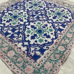 May include: A square, white textile with a repeating blue and teal floral pattern. The center design features large, stylized flowers. The border has a teal floral design with red accents. The textile is likely a scarf or a decorative cloth.