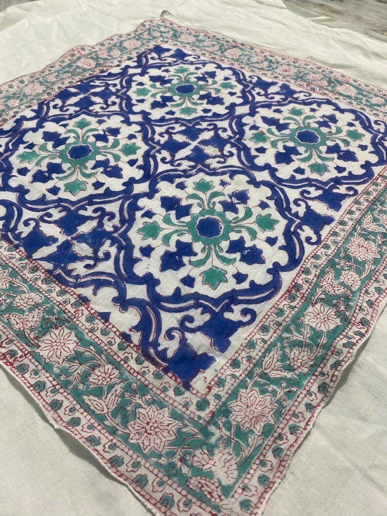 May include: A square, white textile with a repeating blue and teal floral pattern. The center design features large, stylized flowers. The border has a teal floral design with red accents. The textile is likely a scarf or a decorative cloth.
