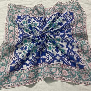May include: Square scarf with a white background and a central blue floral design. The border features a green and pink floral pattern. The scarf is made of a lightweight fabric and is folded.