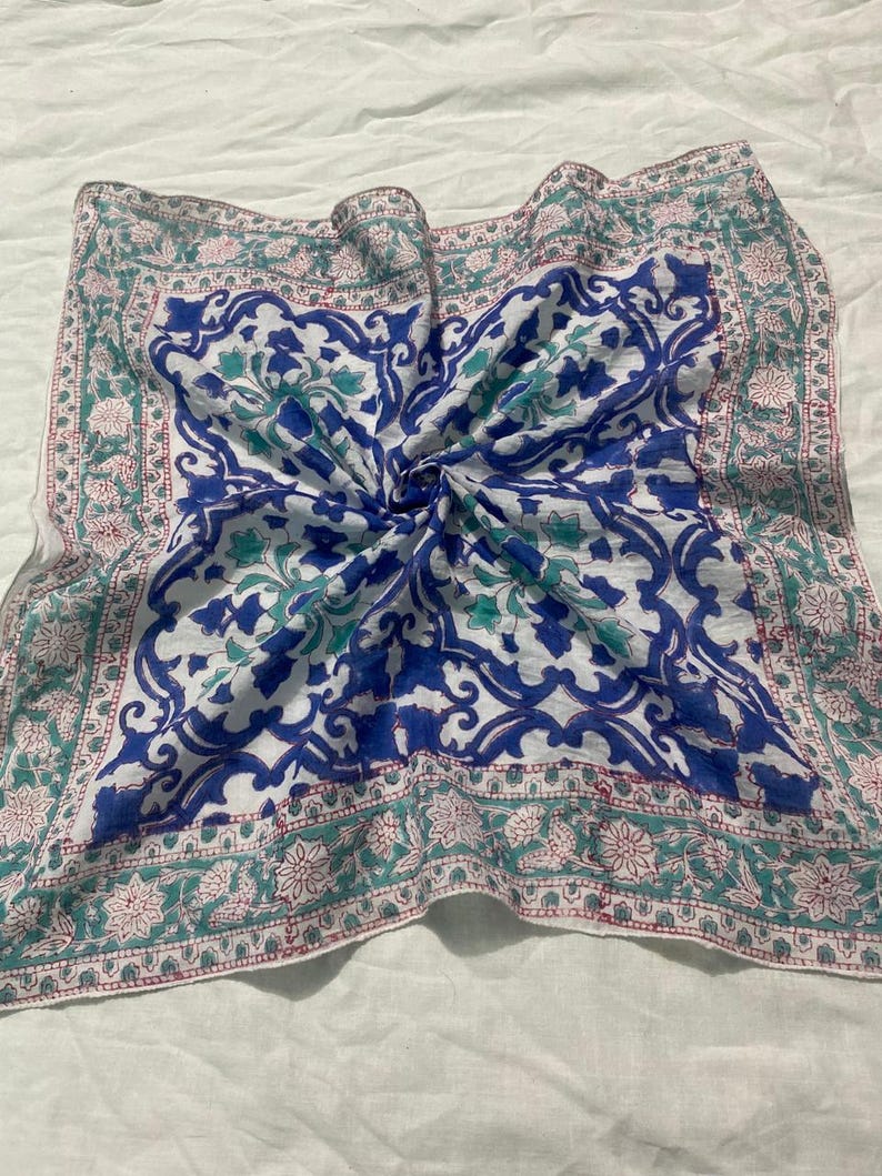 May include: Square scarf with a white background and a central blue floral design. The border features a green and pink floral pattern. The scarf is made of a lightweight fabric and is folded.