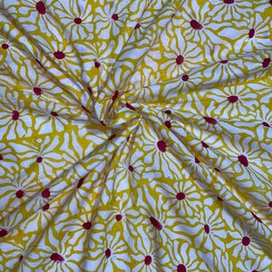 May include: Yellow fabric with a repeating pattern of white daisy-like flowers with red centers. The fabric is wrinkled, suggesting it is a textile. The design is likely for apparel or home decor.